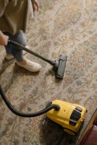 Eine Person saugt mit einem gelben Staubsauger einen Teppich mit floralem Muster.